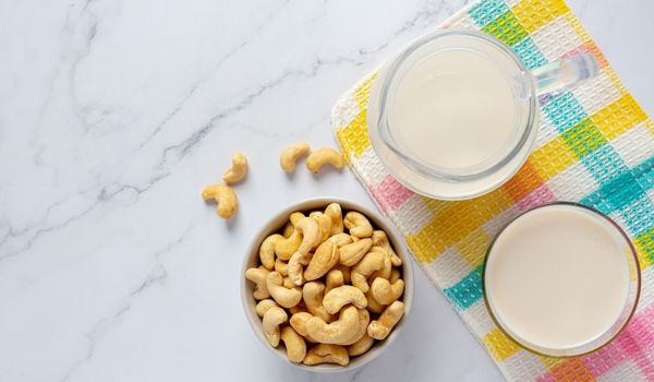 A Bowl of Cashews Lies Next to a Jug, and a Glass of milk Placed Over a Colorful Checkered Table Mat on a Grey Slab