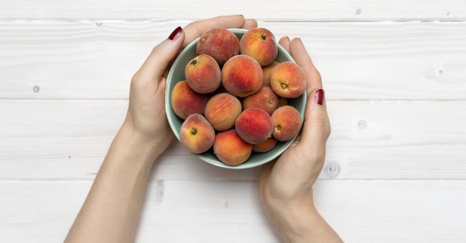 A woman’s hands hold a green bowl full of peaches on a white background