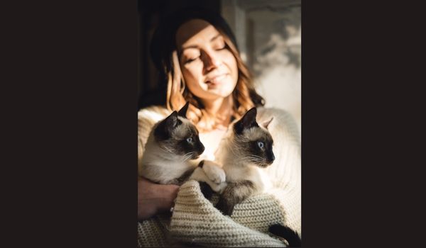 : A woman holds two Siamese kittens while exposing herself to sun rays coming from a window